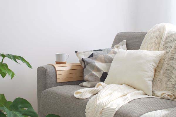 Grey couch with two cushions, a white throw and a cup of tea. Plant in the left hand corner.